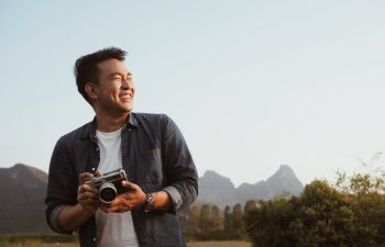 A person smiles while holding a camera outdoors, with mountains and greenery in the background.